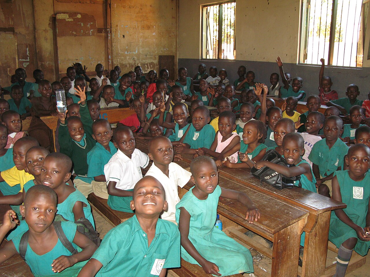 A crowded primary school classroom in Kampala, pupils at worn wooden desks with their hands up