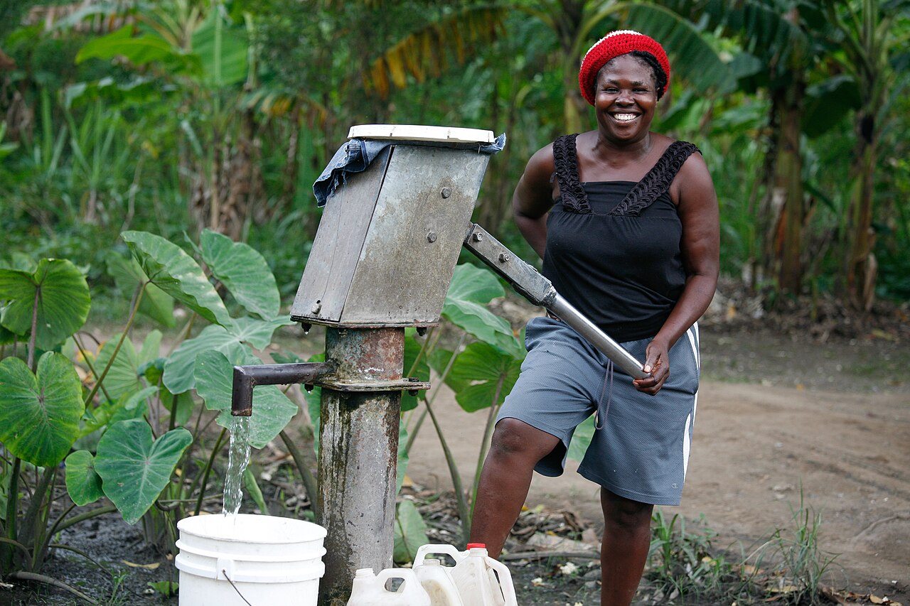 A smiling woman working the handle of a community water pump